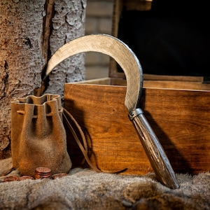 Könnte beinhalten: Eine rustikale Szene mit einer gebogenen Metallsichel mit Holzgriff, einem kleinen Lederbeutel und einer Holzkiste. Mehrere Kupfermünzen sind auf einer fellartigen Oberfläche verstreut, was ein Gefühl von Geschichte erzeugt.