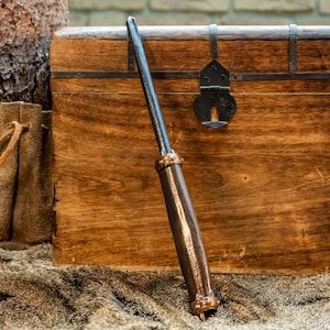 Könnte beinhalten: Ein Metallwerkzeug mit einem langen, schlanken, dunkel gefärbten Schaft und einem dicken, zylindrischen Griff mit metallischem Finish. Der Griff ist an beiden Enden mit kupferfarbenen Bändern akzentuiert, die jeweils kleine, erhabene kreisförmige Details aufweisen.