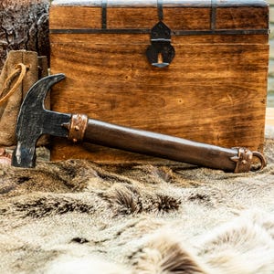 Könnte beinhalten: Ein schwarzer Hammer mit einem dunkelbraunen Holzgriff, der mit kupferfarbenen Metallbändern mit kleinen Nieten an der Ober- und Unterseite des Griffs versehen ist und einen kleinen Metallring am Griffende aufweist, horizontal liegend.
