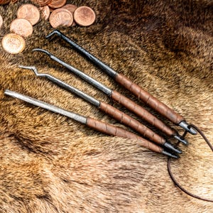 Könnte beinhalten: Ein Set aus vier Metall-Dietrichen mit dunklen, texturierten Stahlschäften und braunen, lederumwickelten Griffen. Jedes Werkzeug hat eine einzigartige Spitzenform, darunter Haken und ein Rechen, und sie sind mit einer dünnen braunen Lederschnur verbunden.