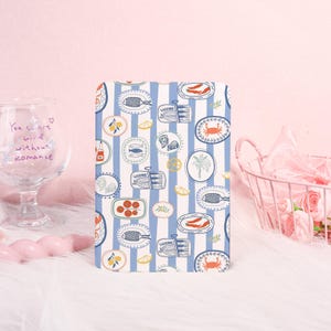 May include: A rectangular notebook with a blue and white striped pattern. The design features illustrations of sardines, lemons, oysters, and crabs. The notebook is on a white surface.