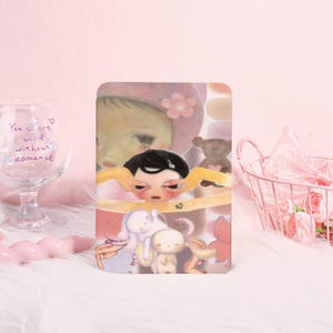 May include: A rectangular art print featuring a whimsical illustration of a child with black hair, surrounded by cartoon figures and a teddy bear. The print has a soft, pastel color palette with pink and beige tones.