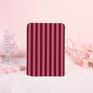 Op de afbeelding: Een rechthoekig object met een verticaal streepjespatroon in tinten bordeauxrood en roze. Het object staat tegen een zachtroze achtergrond met een decoratief glas en een draadmand vol bloemen.