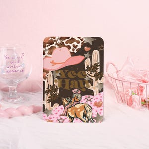 May include: A rectangular notebook with a cowgirl theme. The cover features a pink cowboy hat, cacti, stars, and the words "Yee Haw." A cowgirl rides a horse on the bottom of the cover. The background is a mix of brown, pink, and white.