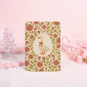May include: A rectangular card featuring a white goose with a red bow, surrounded by a floral pattern of pink flowers and green leaves. The background is a cream color. The card is on a white surface.