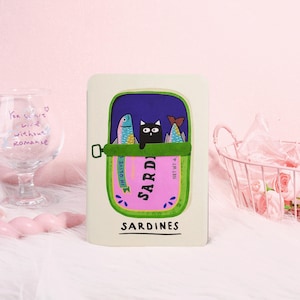 May include: A rectangular, off-white notebook with a whimsical sardine can design. The illustration features a black cat and two fish inside a can labeled "Sardines." The can is pink with green and purple accents, and the word "Sardines" is written at the bottom.