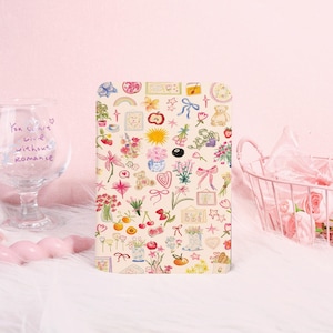 May include: A beige notebook with a colorful pattern of flowers, bows, teddy bears, and other small illustrations. The notebook is rectangular and has a black button closure. The background is a soft pink.