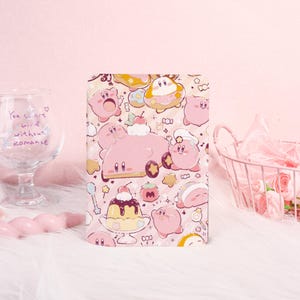 Könnte beinhalten: Ein rosa Notizbuch mit einem sich wiederholenden Muster aus Zeichentrickfiguren und Desserts. Das Cover-Design umfasst verschiedene Kirby-Charaktere, Kuchen und Süßigkeiten. Der Hintergrund ist hellrosa und die Gesamtästhetik ist niedlich und verspielt.