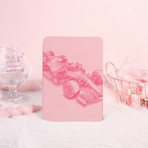 Op de afbeelding: Een roze notitieboek met een gedetailleerde illustratie van een Formule 1-raceauto. De auto is weergegeven in een monochrome roze kleur, met zichtbare details van de wielen, het chassis en de aerodynamische elementen. Het notitieboek heeft afgeronde hoeken.