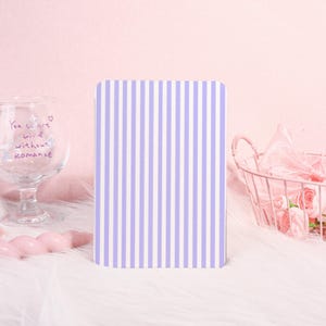 May include: A rectangular notebook with a vertical striped pattern in lavender and white. The notebook is the focal point, set against a soft, pink background with a glass and a wire basket.