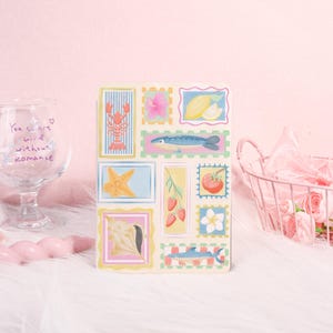 May include: A decorative notebook featuring colorful illustrations of various sea creatures, fruits, and flowers within postage stamp-like frames. The notebook has a cream-colored background and is set against a soft pink backdrop.
