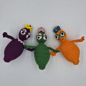 May include: Three handmade crochet dolls in purple, green, and orange. Each doll has a flower crown and large eyes. The dolls are holding hands, showcasing a whimsical design. The dolls are approximately 6 inches tall.