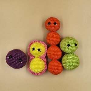 May include: A collection of handmade crochet toys in various colors. The toys include a purple sphere, a yellow and pink figure, a stack of orange spheres, and two green spheres. Each toy has black button eyes.