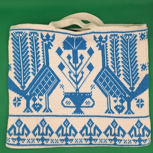May include: A white crocheted tote bag with a blue geometric pattern featuring stylized birds and floral motifs. The bag has a rectangular shape with a handle, perfect for carrying items. The design is symmetrical and detailed.