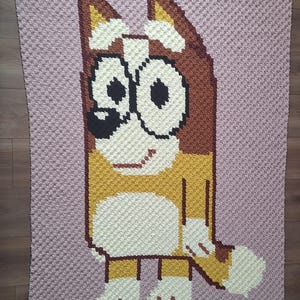 May include: A handmade crochet blanket featuring a cartoon dog character in shades of brown, yellow, white, and black, set against a light purple background. The blanket is crafted with a textured stitch, showcasing the character's features.