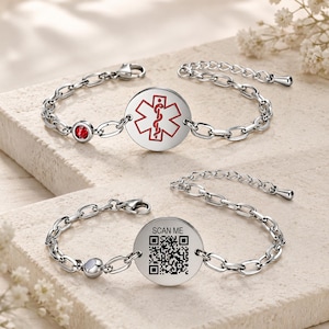 Puede incluir: Dos pulseras de alerta médica plateadas. Una presenta una gema roja y un símbolo médico rojo sobre un disco blanco. La otra tiene un código QR y el texto "SCAN ME" en un disco blanco. Ambas tienen eslabones de cadena.