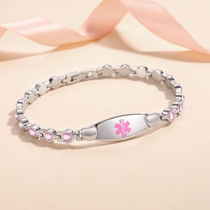 Puede incluir: Una pulsera de alerta médica plateada con un símbolo médico rosa. La pulsera presenta gemas rosas y detalles en forma de corazón. El símbolo médico está centrado en una placa rectangular.