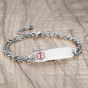 Könnte beinhalten: Ein silberfarbenes medizinisches ID-Armband mit einem Kettendesign. Die rechteckige Platte zeigt ein rotes Medizinsymbol. Das Armband wird auf einer Holzoberfläche präsentiert.