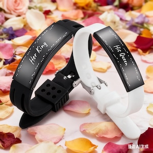 Puede incluir: Dos pulseras a juego, una negra y otra blanca, con las frases "Her King" y "His Queen" inscritas. Las pulseras tienen un diseño elegante y moderno con una correa de silicona y un cierre metálico. El fondo es una mezcla de pétalos de flores coloridos.