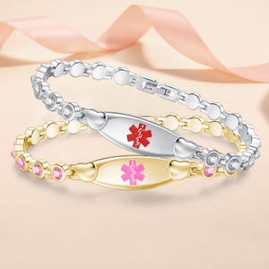 Könnte beinhalten: Zwei medizinische Notfallarmbänder mit einer zentralen ovalen Plakette, die ein rotes oder rosa Symbol des Sterns des Lebens zeigt. Das silberfarbene Armband hat klare Steine, das goldfarbene rosa Steine. Beide haben herzförmige Akzente an der Plakette.