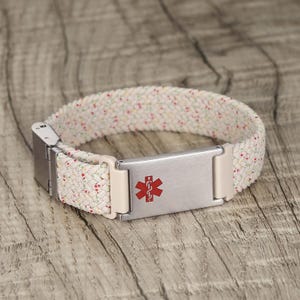 Op de afbeelding: Crème medische ID-armband met een gevlochten band met rode, gele en groene spikkels. De zilveren metalen plaat heeft een rood Star of Life-symbool. De armband heeft een veilige sluiting.