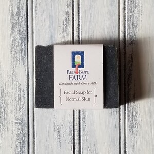 Puede incluir: Una pastilla de jabón facial gris oscuro para piel normal, con una etiqueta blanca que dice "Red Rope Farm Handmade with Goat's Milk". El jabón es rectangular y está sobre una superficie de madera blanca y gris.