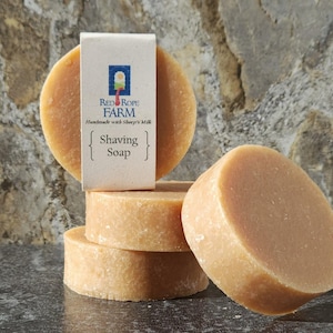 Op de afbeelding: Vier ronde, lichtbruine scheerzeepjes zijn op elkaar gestapeld, met een label met de tekst "Red Rope Farm Handmade with Sheep's Milk Shaving Soap". De zepen liggen op een donker, getextureerd oppervlak, met een stenen achtergrond.