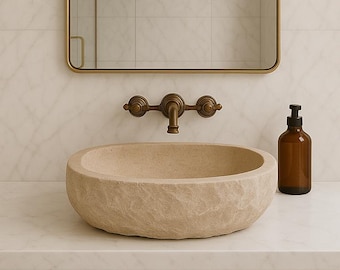 Puede incluir: Un lavabo de piedra beige de forma redonda con una superficie exterior texturizada y rugosa. Se combina con un grifo de pared de latón antiguo con dos manijas y un caño central, situado debajo de un espejo cuadrado con un marco fino de latón.