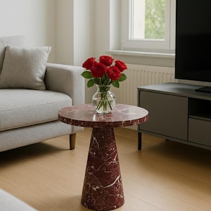 May include: A round side table with a red marble top and a matching conical base. A clear glass vase holds a bouquet of red roses. The table is in a living room setting with a gray sofa and a TV stand.