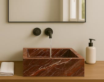 Op de afbeelding: Een moderne badkamer wastafel van rood marmer met witte adering. De rechthoekige wastafel staat op een houten wastafelmeubel. Een zwarte kraan en zeepdispenser zijn zichtbaar. Een witte handdoek ligt op het wastafelmeubel.