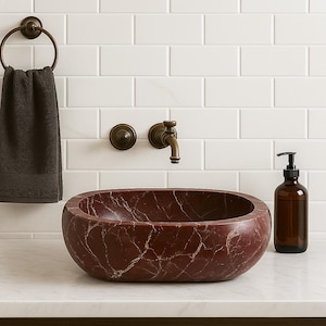 Può includere: Un lavabo da bagno in marmo rosso con venature bianche. Un asciugamano marrone scuro è appeso a un anello portasciugamani in bronzo. Sono visibili anche un rubinetto e un dosatore di sapone in bronzo.