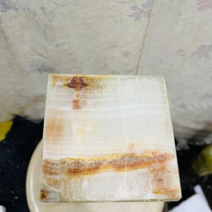 Puede incluir: Un posavasos cuadrado de &oacute;nix blanco con vetas marrones y naranjas. El posavasos est&aacute; hecho de piedra natural y tiene una superficie lisa y pulida.