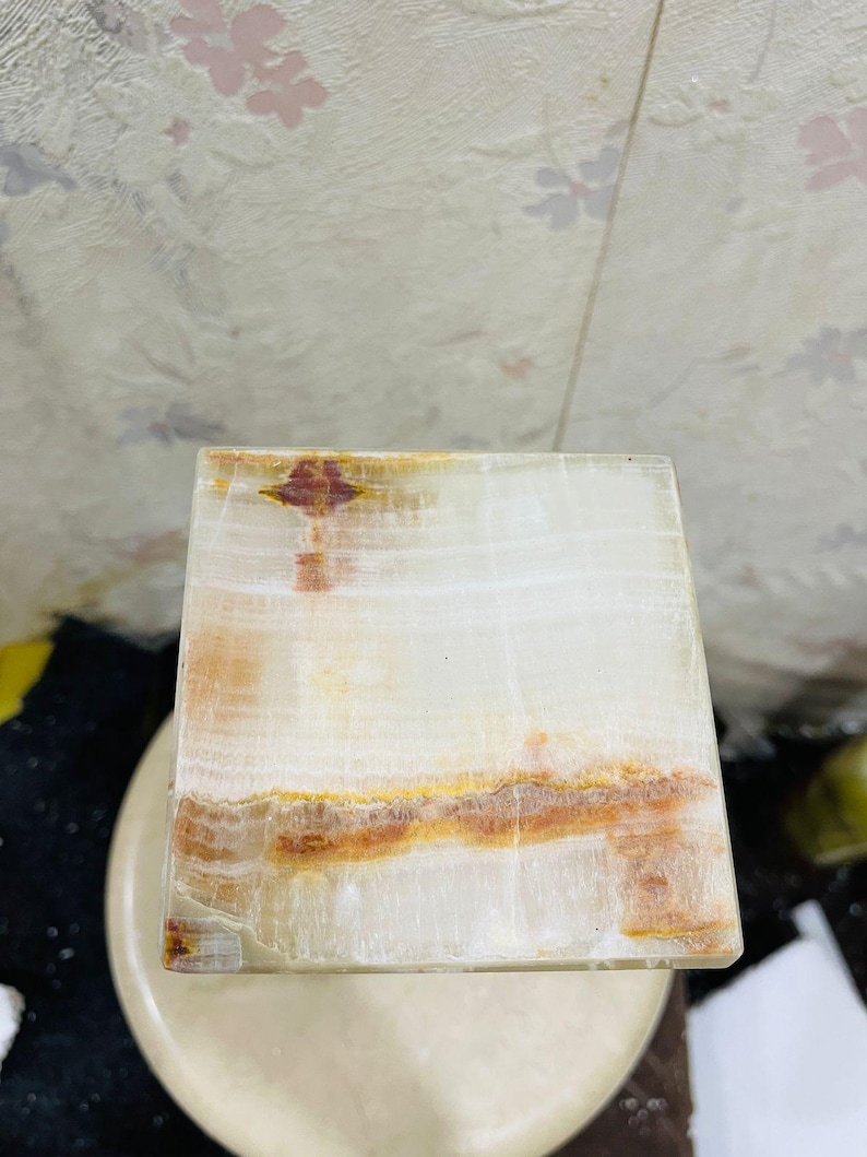 Puede incluir: Un posavasos cuadrado de &oacute;nix blanco con vetas marrones y naranjas. El posavasos est&aacute; hecho de piedra natural y tiene una superficie lisa y pulida.