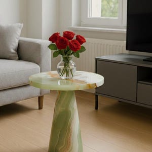 Pode incluir: Uma mesa lateral redonda com um tampo de mármore verde e branco e uma base cónica. Um vaso de vidro com rosas vermelhas está sobre a mesa. A mesa está numa sala de estar com um sofá cinzento e um armário cinzento escuro.