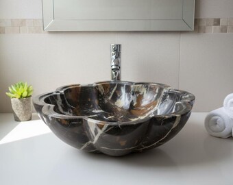 May include: A decorative, flower-shaped bathroom sink made of dark marble with brown and white veining. The sink is set on a white countertop, with a chrome faucet, a small potted plant, and rolled white towels in the background.