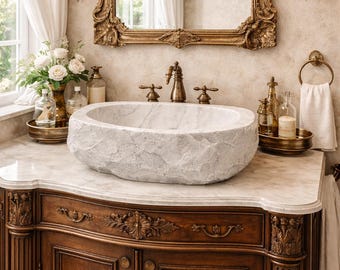 Puede incluir: Un lujoso mueble de baño con un lavabo ovalado con aspecto de piedra. El mueble de madera oscura presenta tallas ornamentadas y una encimera de mármol. Un espejo con marco dorado y apliques con detalles de cristal completan el elegante diseño.