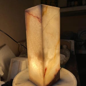 Puede incluir: Una l&aacute;mpara rectangular de alabastro blanco con vetas marrones y amarillas. La l&aacute;mpara est&aacute; encendida y proyecta un brillo c&aacute;lido.