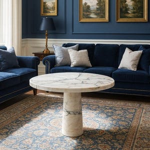 May include: A round marble coffee table with a white and gray patterned top and a cylindrical base. The table sits on a patterned rug in a room with blue velvet sofas and framed landscape paintings.