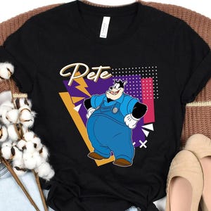 May include: Black t-shirt featuring a cartoon illustration of Pete, with the name "Pete" in a script font. Pete is depicted in blue overalls and black gloves, set against a geometric background with purple, pink, and yellow accents. The shirt is displayed on a brown sweater.