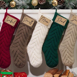 May include: Five cable-knit Christmas stockings in red, brown, white, green, and tan, each with a wooden name tag. Names include Isabel, Daisy, Mom, Dad, and Jacob. Festive holiday decor with pine boughs and ornaments.
