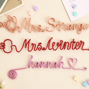 Könnte beinhalten: Drei Draht-Namensschilder: "Mrs. Strange" in Roségold, "Mrs. Winter" in Rot und "hannah" in Pink mit Herz. Dekorative Drahtkunst für Heimdekoration oder Geschenke.