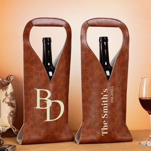 May include: Two brown leather wine bottle carriers with handles, each holding a wine bottle. One carrier has the initials "BD", the other says "The Smith's est.2025". A wine glass with red wine is on the right.