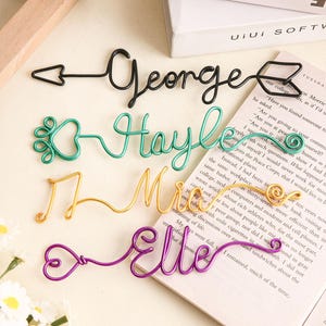 以下が含まれることがあります： 黒、緑、金、紫のワイヤーネームブックマーク。「George」、「Hayle」、「Mrs」、「Elle」という名前が筆記体で書かれています。各ブックマークは、矢印、ハート、音符など、ユニークなデザインになっています。