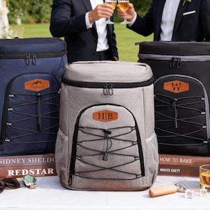 May include: Three insulated cooler backpacks in navy blue, gray, and black, each with a front pocket and a brown leather patch with custom initials or a name. The backpacks have black bungee cords and zippers. The scene includes beer bottles and glasses.