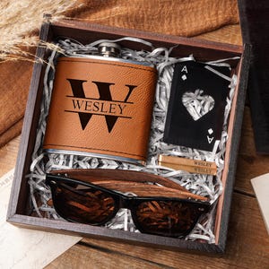 May include: A wooden gift box containing a brown leather flask with "WESLEY" engraved, a black Ace of Hearts playing card, two small wooden items with "WESLEY" text, and wooden sunglasses. The box is filled with shredded paper.