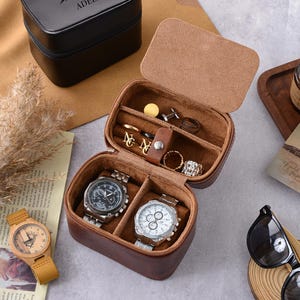 Puede incluir: Una caja de joyería de viaje de cuero marrón con una tapa abierta, que revela compartimentos para anillos, gemelos y dos relojes. También se ve un reloj de madera con una correa amarilla y una caja negra. Otros accesorios incluyen una vela y gafas de sol.