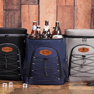 Könnte beinhalten: Drei isolierte Rucksackkühler in Schwarz, Marineblau und Grau, jeweils mit einer Vordertasche und einem personalisierten Kunstlederaufnäher. Der marineblaue Kühler ist offen und mit Bierflaschen gefüllt. Die anderen sind geschlossen.