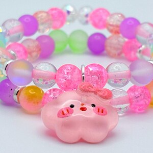 May include: A colorful beaded bracelet with a pink cloud charm. The bracelet features a variety of beads in shades of pink, purple, green, and clear. The cloud charm has a cute face and bow.