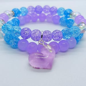 May include: A double-strand beaded bracelet featuring a mix of round crackle-textured and smooth beads in shades of light purple and bright blue. The bracelet includes iridescent clear accent beads and a central purple heart-shaped charm.