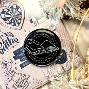 Könnte beinhalten: Eine transparente Handyhülle mit einem hellrosa und weißen Design mit einem herzförmigen Dartboard, Blumen und den Worten "Just Breathe". Ein schwarzer Anstecker mit dem Text "Antisocial Book Club" und einem offenen Buch ist ebenfalls sichtbar.